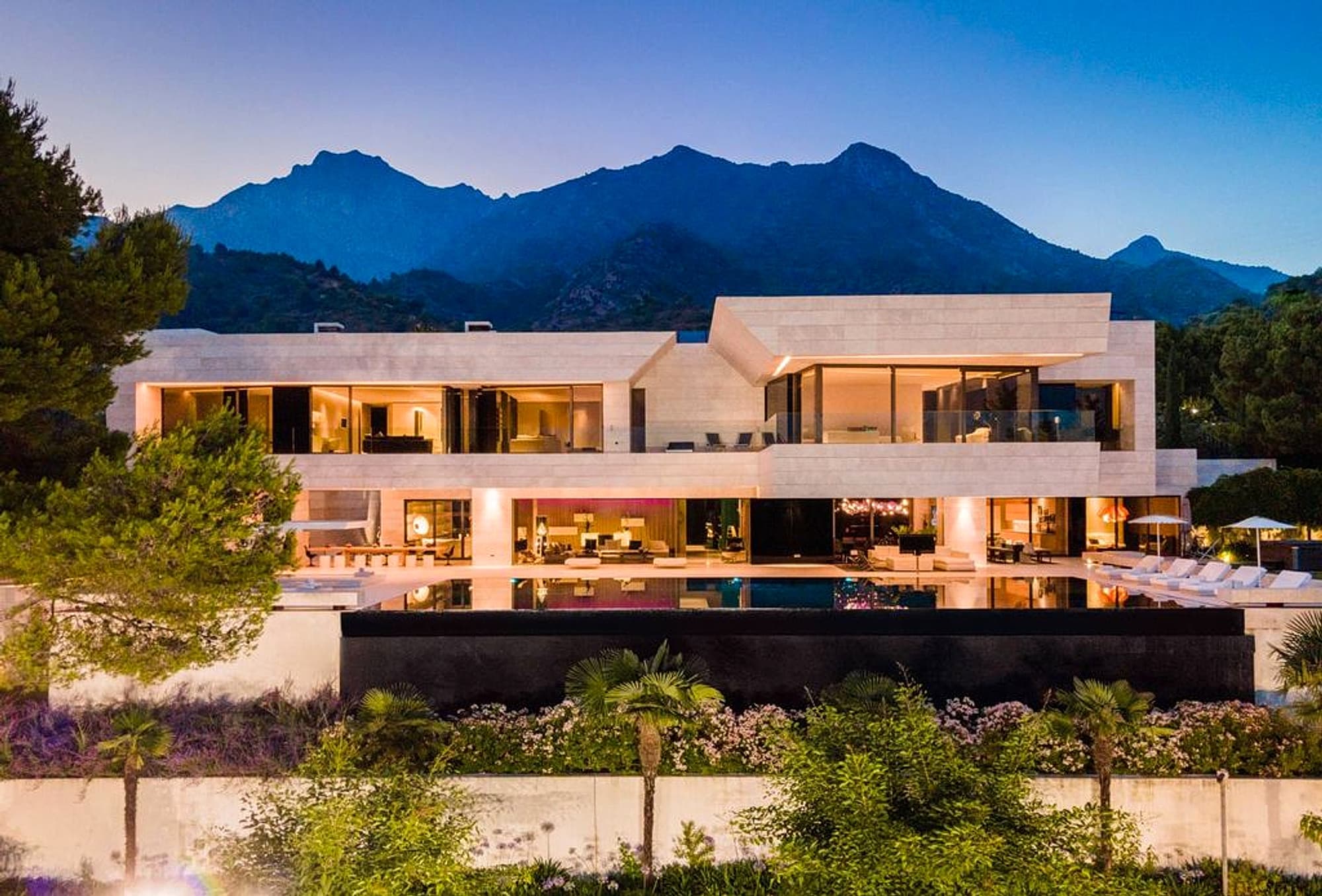 Modern hillside house with pool, glass terraces, and mountain backdrop at dusk